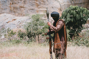 Fototapeta premium Outdoor military concept. Armed man in desert field with rifle and rocket launcher, masked warrior standing alert