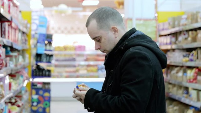Careful male shopper reading nutrition label while standing in supermarket aisle, evaluating product details and making thoughtful purchasing decision