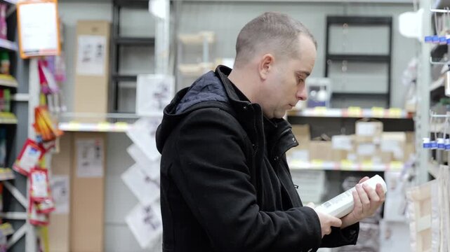 Young male shopper examining product label in grocery store aisle, comparing options while wearing winter jacket and selecting item carefully