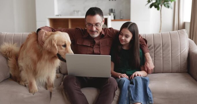 Middle age Caucasian father sitting on couch with daughter, spending leisure time in living room, stroking golden retriever dog, watching movie on laptop. Family indoors relaxation on weekend at home