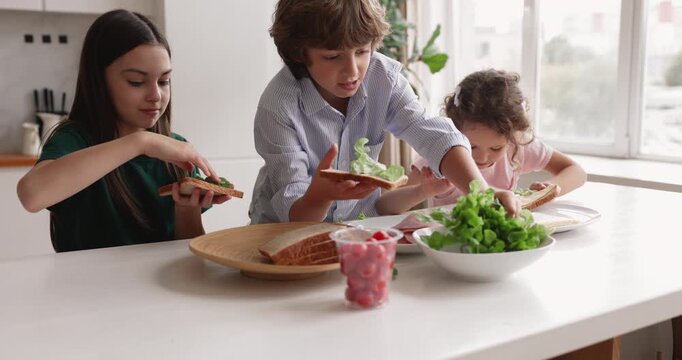 Three Caucasian sibling children making fresh sandwiches with bread, lettuce and meat slices in domestic kitchen during breakfast at home. Healthy eating habit from childhood, family cooking concept