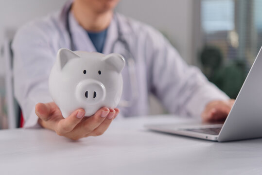 doctor holding white piggy bank medical clinic desk laptop healthcare costs health insurance finance saving money hospital budget investment clinic financial planning wealth medicine economy care