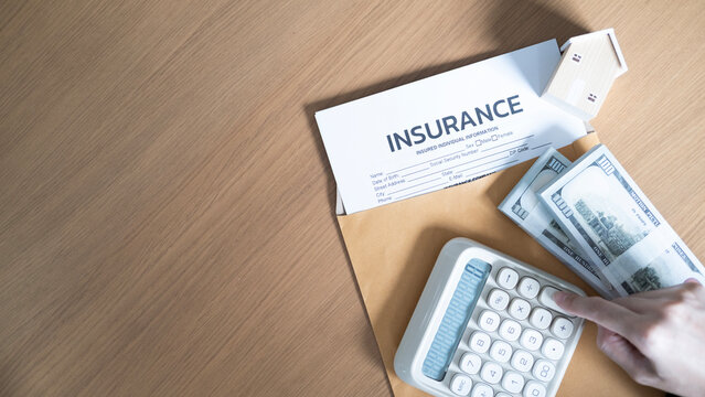 Home insurance and mortgage planning concept with a calculator, insurance form, wooden house model, cash bundles, and copy space on a wooden table.