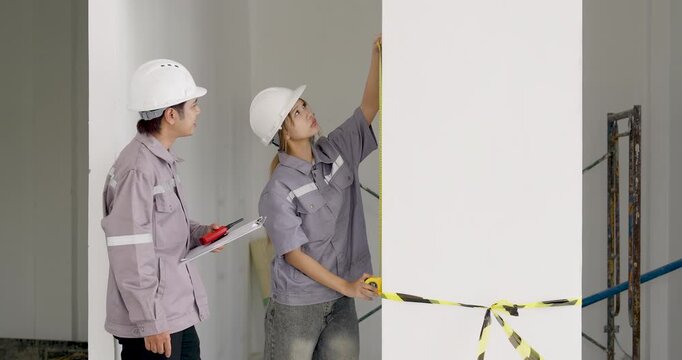 Asian female engineer checks clipboard while male contractor measures wall section, supporting construction document review process, safety planning, quality inspection, progress tracking