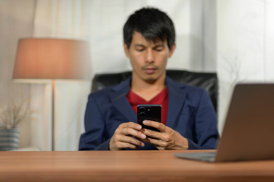 Young man using smartphone at office desk with laptop and lamp in background