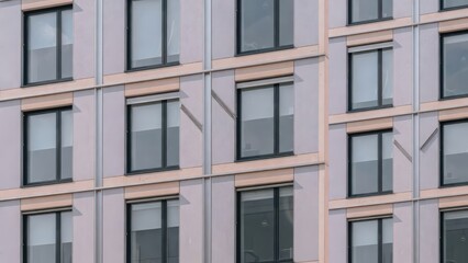 Fototapeta premium A view of the repetitive geometric layout of a tall residential tower featuring symmetrical glass window panes arranged vertically across a colorful gray and peach exterior facade in a dense city