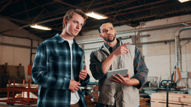 Men, tablet and meeting in lumber workshop, carpenter and apprentice with discussion for advice in woodwork. Point, talk and supervisor with trainee, tech and team guidelines for carpentry planning