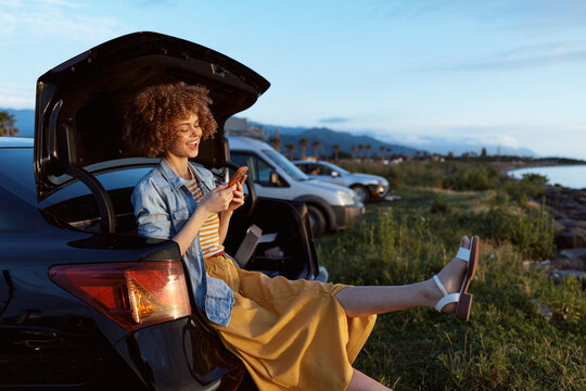 Woman with curly hair smiles while sitting on a car trunk and using a smartphone in an outdoor sunset scene, highlighting lifestyle travel outdoor sunset, casual relaxation and warm mood. Soft