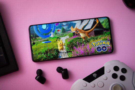 Dhaka, Bangladesh- 23 March 2025: Smartphone displaying Pok&eacute;mon GO game on pink surface with earphones and controller nearby.