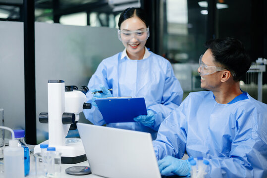 Two scientists collaborating in a modern biotechnology laboratory using laptop and microscope for biomedical research, healthcare innovation and pharmaceutical development.