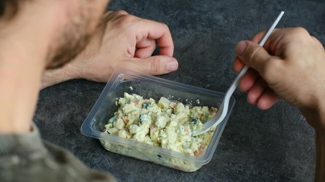 Man eating pre packaged salad from transparent plastic container bought at supermarket, with disposable fork. Convenience meal unhealthy processed food. Man eating pre packaged salad from supermarket 