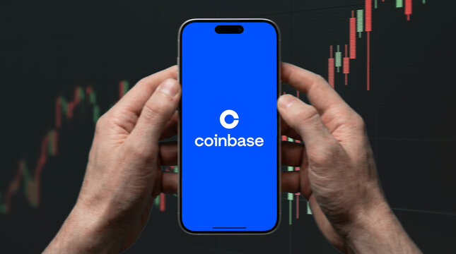 Coinbase cryptocurrency exchange app logo on iPhone screen held with both hands against candlestick chart background