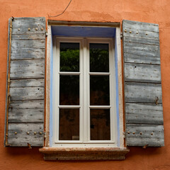 Obraz premium A classic Mediterranean window with blue wooden shutters set against an ochre-colored exterior wall. Rustic, weathered wooden shutters in a faded gray-blue, typical of Provence in France