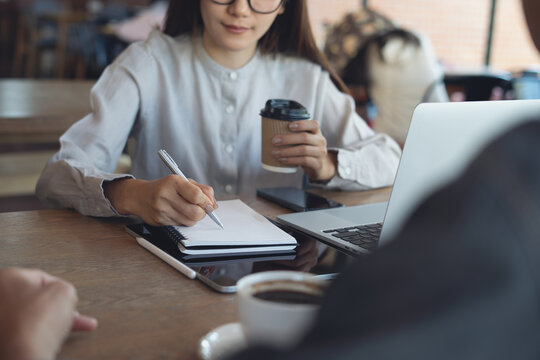 Businesswoman writing notes in notebook, holding coffee cup with laptop at table, working at cefe, modern workspace, meeting or remote work concept