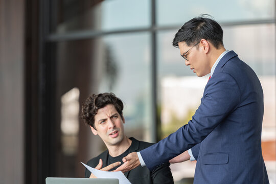 Stressed young businessman holding head in hand receiving criticism by manager after discussing document error on laptop for work mistake at outdoor office business pressure leadership feedback.