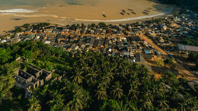 Aerial view of Fort Batenstein ruins overlooking the coastal village of Butre with traditional houses, palm trees, and fishing boats on the sandy beach in Butre, Western Region, Ghana.