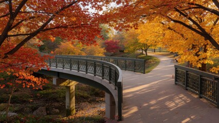 Obraz premium A picturesque walking path leads across a curved metal bridge through a tranquil park filled with vibrant red and golden maple trees during a sunny afternoon at the peak of the autumn season chan