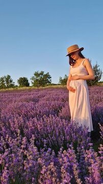 A beautiful pregnant woman smiles and poses in a vast lavender field. Wearing a pink dress and straw hat, she cradles her baby bump, enjoying the endless rows of purple lavandula flowers at sunset