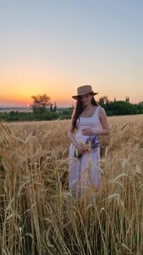 A beautiful pregnant woman stands in a golden wheat field at sunset. Wearing a straw hat and a light pink sundress, she holds a lavender bouquet and cradles her baby bump, looking down peacefully