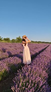 A beautiful pregnant woman smiles and poses in a vast lavender field. Wearing a pink dress and straw hat, she cradles her baby bump, enjoying the endless rows of purple lavandula flowers at sunset
