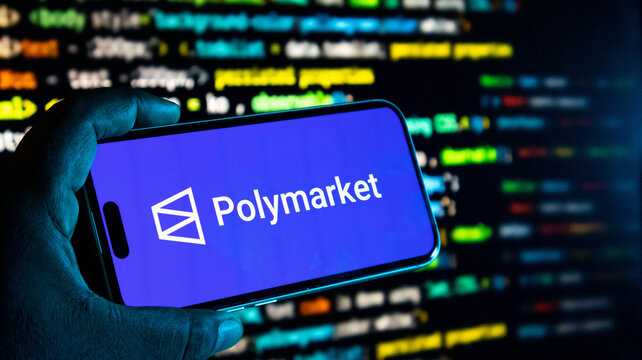 Hand holding a smartphone displaying the polymarket logo against a background of blurred computer code on a screen