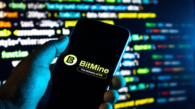 Closeup of a hand holding a smartphone displaying the bitmine logo against a background of blurred computer code