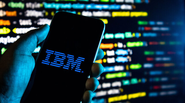 Hand holding a smartphone with the ibm logo displayed on the screen against a background of computer code