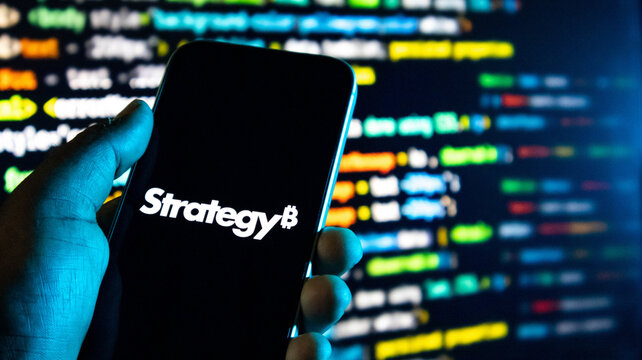 Hand holding a smartphone displaying a bitcoin strategy logo against a background of computer code