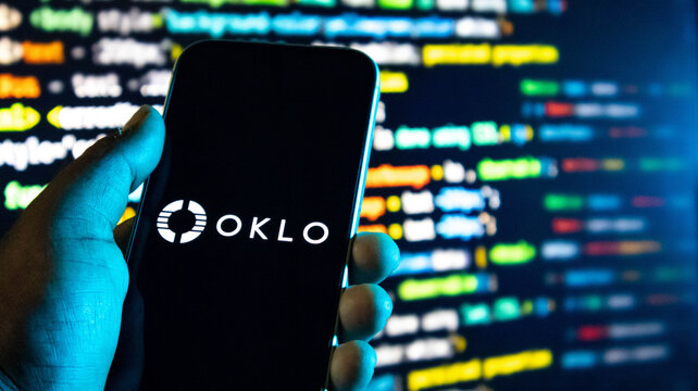 Closeup of a hand holding a smartphone displaying the oklo logo against a background of blurred computer code