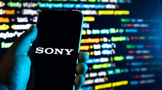 Hand holding a smartphone displaying the sony logo against a background of blurred computer code on a monitor screen