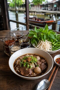 raditional Thai boat noodles served with fresh vegetables on wooden table, floating market background