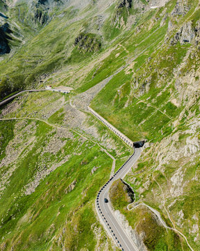 Aerial view of the Great St. Bernard Pass winding through the lush green alpine slopes with a car driving along the mountain road in Saint-Rhemy, Valle d'Aosta, Italy.