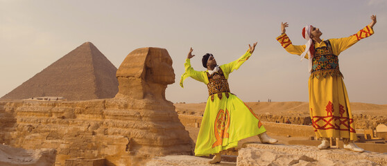 Fototapeta premium Two tanoura performers in vivid embroidered robes raise arms in joyful dance before the Great Sphinx and Khafre Pyramid at Giza plateau Egypt ideal