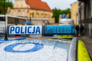Close-up of the roof of a marked Polish police patrol car featuring a blue emergency light bar with the sign POLICJA, covered in raindrops, against a blurred urban architectural background © Adam