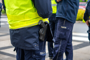 Close-up of Polish police officers' uniforms, featuring a holster with a service weapon at the belt, dark blue trousers with the word POLICJA, a high-visibility yellow vest, and tactical equipment dur © Adam