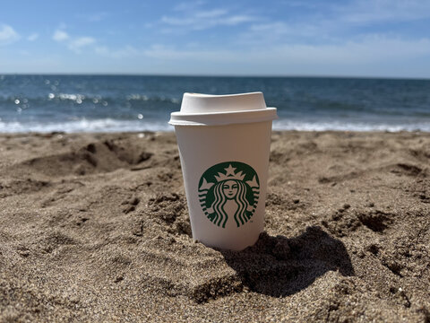 ANDALUSIA, SPAIN - APRIL 22, 2025: relaxed starbucks coffee cup resting in sand on beach with ocean view ground-level perspective summery vacation, copy space.