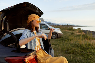 Naklejka premium Cheerful woman outdoors, sitting at a car trunk, sunlit moment, casual dress and scarf, carefree lifestyle, breezy afternoon, scenic coastline background