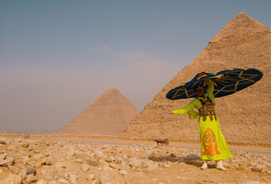 Festive egyptian folk performer in neon yellow embroidered costume balances large decorative umbrella before the iconic ancient pyramids on the rocky