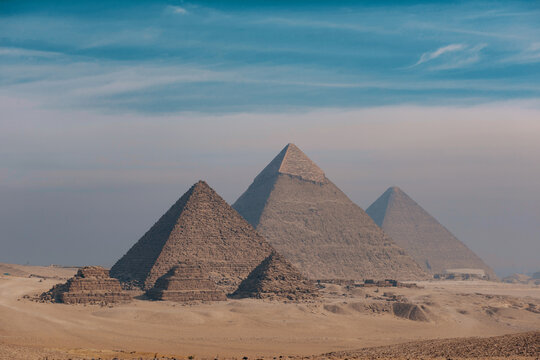 Panoramic view of the three Pyramids of Giza rising in staggered formation above the sandy Giza plateau under a hazy blue sky ideal for travel