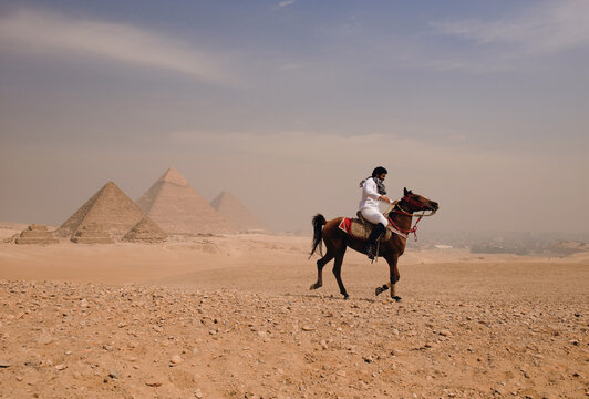 Dramatic wide shot of an Arab horseman in white traditional garment galloping confidently across Giza desert plateau with iconic Great Pyramids