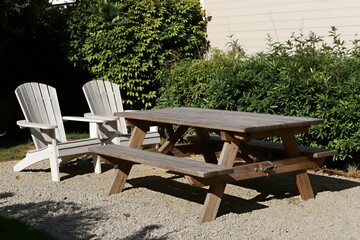 Fototapeta premium Wooden picnic table with benches, Adirondack chairs sits on gravel path. Rich green bushes, light colored building wall form background. Outdoor seating area offers relaxation, natural setting.