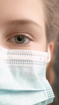 A girl is wearing a blue surgical mask, covering her mouth. Her eye is visible, showing calm determination and attention to health safety.