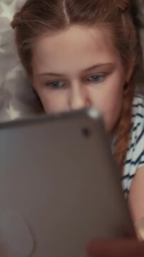 A girl is focused on a tablet device, engaging with its content. The girl appears relaxed as she uses the tablet in a comfortable setting.