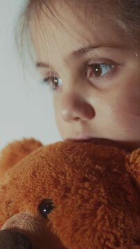 A girl holds a brown teddy bear close to her face. The girl's expression is pensive, as she gazes off into the distance, with gentle eyes reflecting emotion.