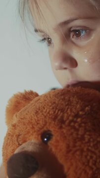 A girl holds a teddy bear while looking down. The girl appears sad, with tears on her cheeks, emphasizing her emotional connection to the teddy bear.