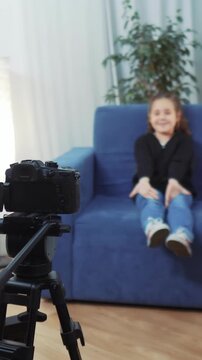 A girl is sitting on a blue couch, looking at a camera. The camera is placed on a tripod, focused on the girl as she smiles in the cozy room.