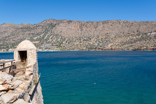 Historic stone watchtower booth stands on fortress wall by bright blue sea water with distant mountains under clear sky on Spinalonga island Crete Greece.