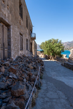 Old ruined houses with wooden frames and stone walls stand along empty street on historic Spinalonga island under clear sky in Crete Greece.