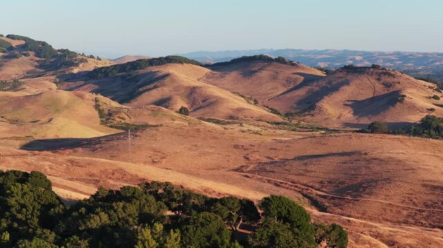 Telephoto helix pulling back from oak trees on a golden hillside at sunset, revealing rolling hills, distant mountain ranges and transmission lines in warm Napa County light.