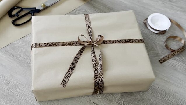 Gift-wrapped package with brown paper and leopard print ribbon, surrounded by scissors, wrapping paper roll, and tape on a wooden surface
